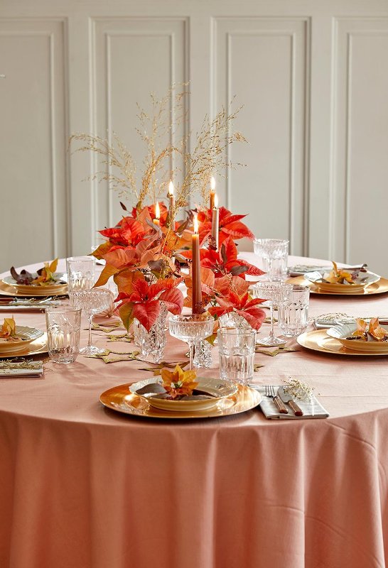 a bold and chic wedding tablescape with a pink tablecloth, gold chargers, a large centerpiece of poinsettias in glasses and rust-colored candles