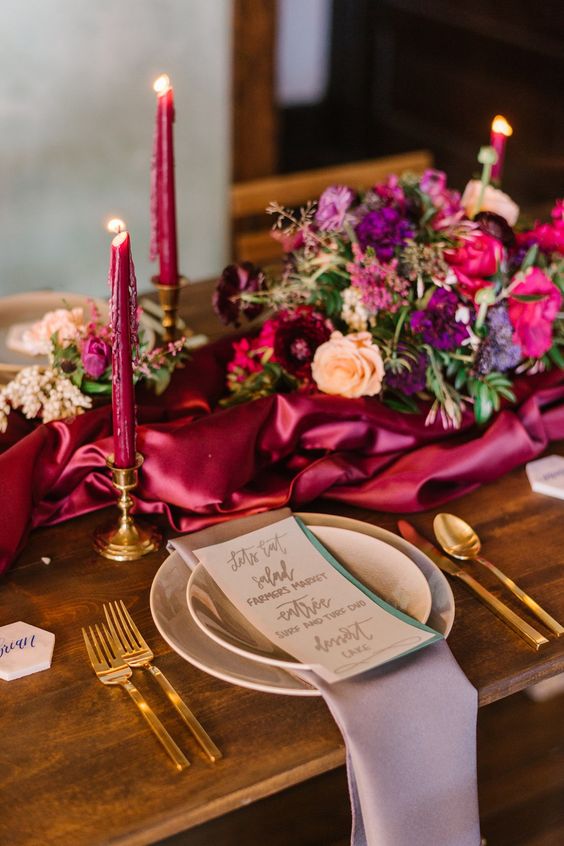 a chic wedding tablescape with a magenta runner and matching candles, bright blooms and greenery, neutral plates and a lilac napkin