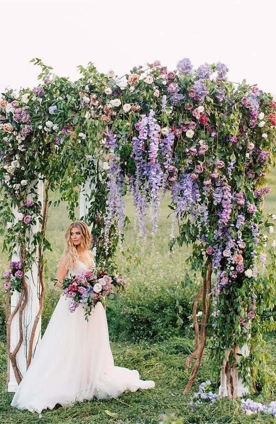 a very natural wedding arch with much greenery, purple and light pink blooms and some neutral fabric is wow