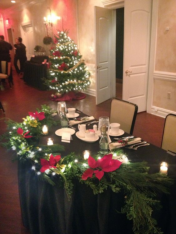 a sweetheart wedding tablescape with a black tablecloth, candles, an evergreen garland with lights and red poinsettias