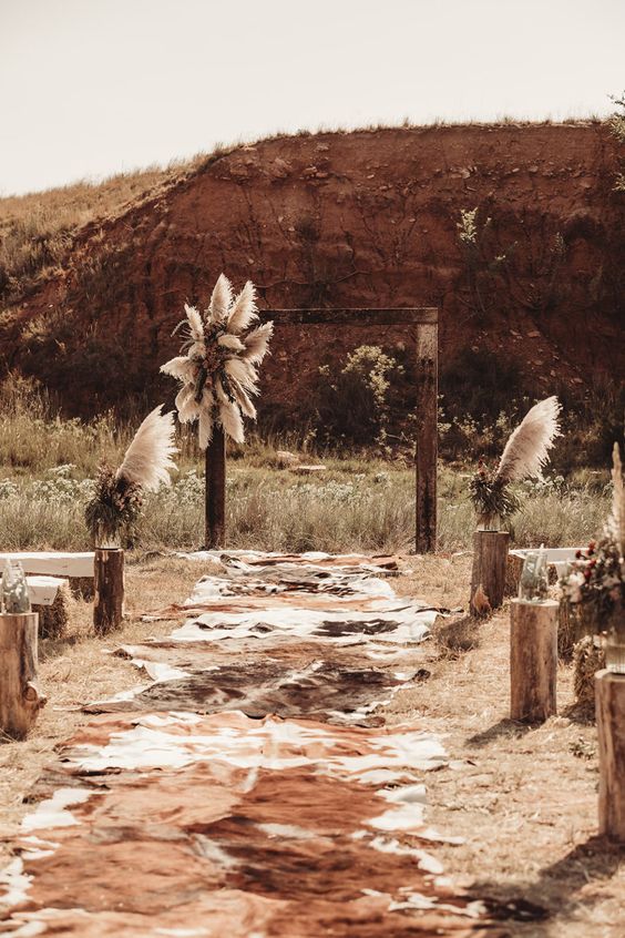 a boho Western wedding ceremony space with an arch decorated with pampas grass and greenery, cowhide rugs on the aisle