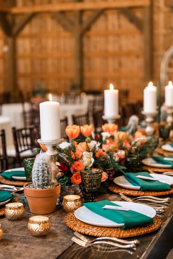 a Western wedding tablescape with orange blooms, potted cacti, candles, woven placemats, emerald napkins is amazing