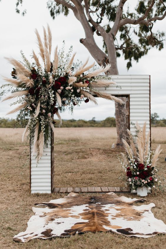 a Western wedding arch done with blusha nd burgundy blooms, greenery and pampas grass plus a cowhide rug