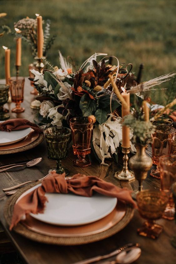 a bright mid-Western wedding table setting with dark blooms and greenery, colored glasses and amber candles, rust and orange touches