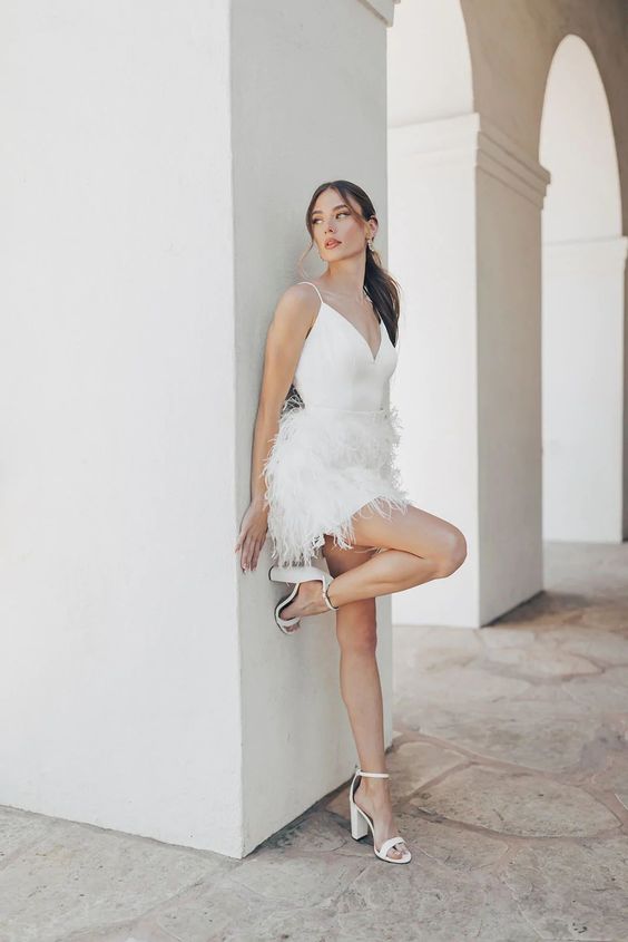 a mini white wedding dress with spaghetti straps and a V-neckline plus a feather skirt and block heels are a trendy wedding outfit