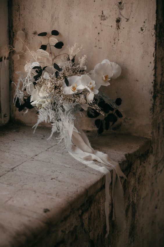 a beautiful black and white wedding bouquet of white orchids and lunaria, black leaves and dried grasses and neutral ribbons