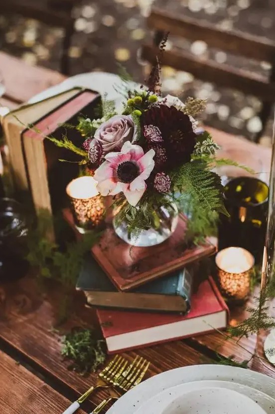 a beautiful dark enchanted forest wedding centerpiece of a stack of books, candles, a dark floral centerpiece and fern