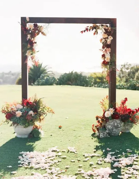 a bold rustic fall wedding arch decorated with burgundy and blush dahlias, greenery and bold foliage, with matching arrangements in vases at the base