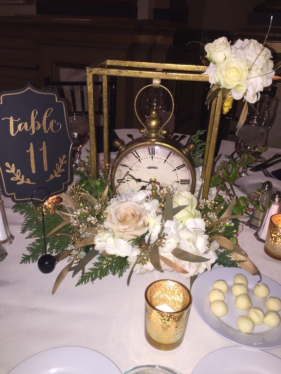 a chic NYE wedding centerpiece of white blooms, greenery, gilded leaves, aframe with a suspended clock and a table number