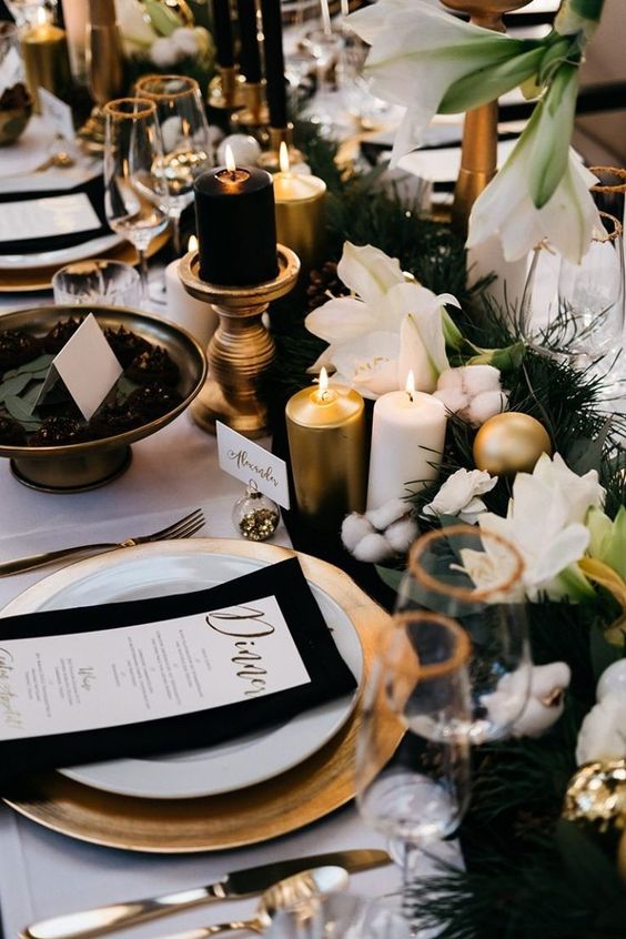 a chic NYE wedding tablescape with a greenery runner, white blooms, cotton and gold ornaments, pillar candles, gold chargers and black napkins