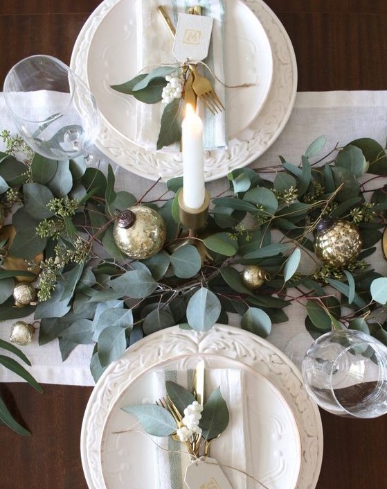 a festive table runner with frsh greenery and gold ornaments plus tall candles make the tablescape chic