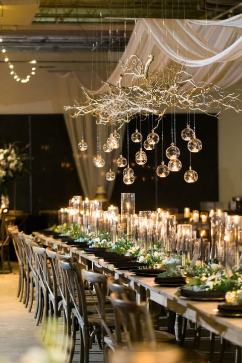 a gorgeous NYE wedding tablescape with greenery and white blooms, black plates, a gold branch with gold candles hanging over the table