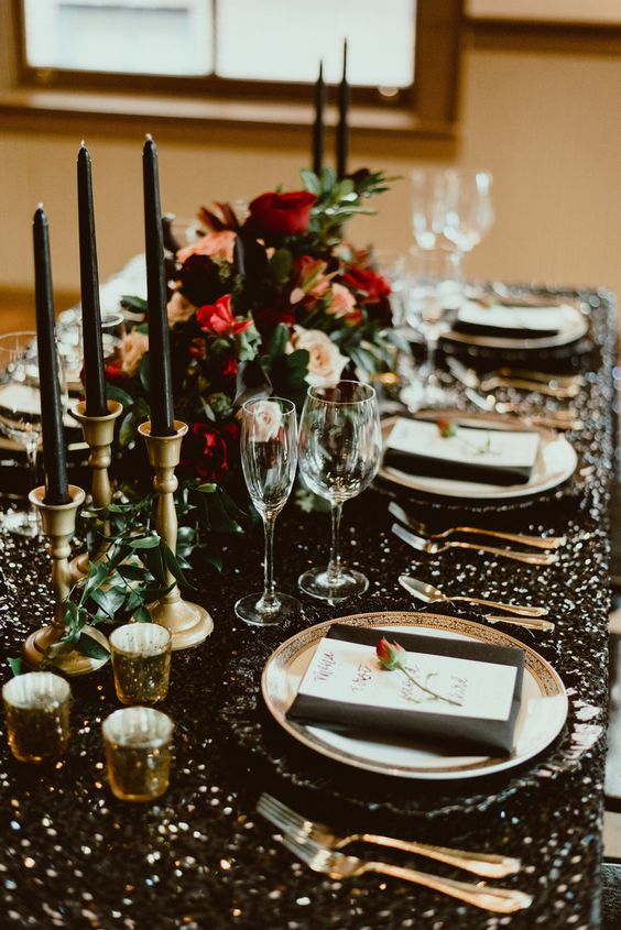a gorgeous black, gold and red NYE wedding tablescape with black napkins and tall and thin candles, gold cutlery