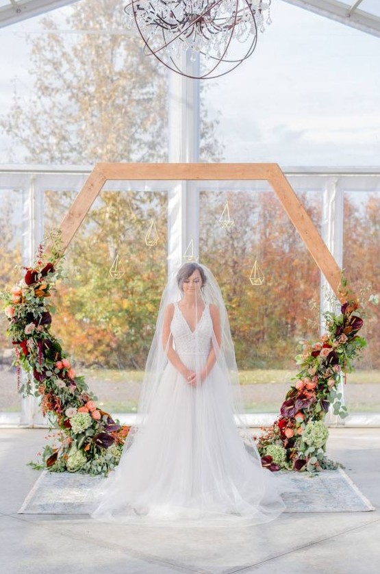 a hexagon wedding arch with greenery and dark and bold flowers and candle lanterns hanging for a fall wedding