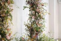 a jaw-dropping garden wedding arch covered with greenery, yellow, blush and white blooms and some arrangements at the base looks fantastic