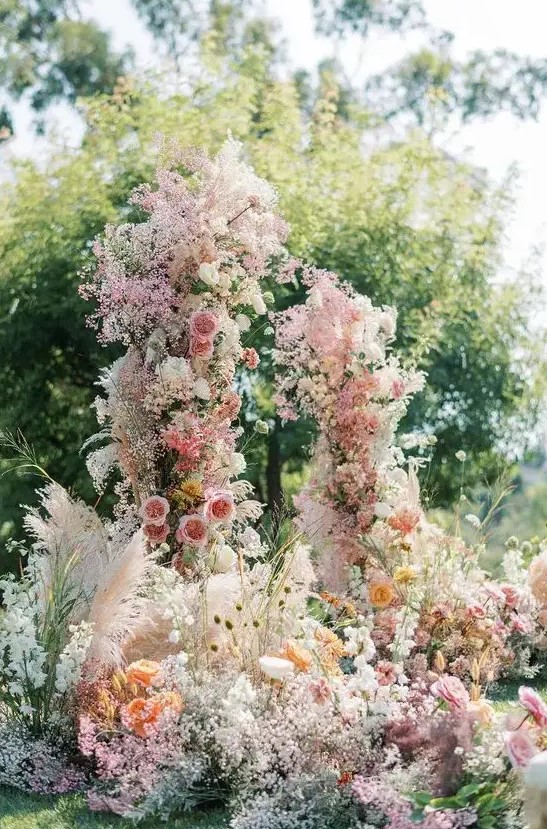 a jaw-dropping wedding arch with pink, orange and white blooms, dyed baby's breath and pampas grass is gorgeous for a garden wedding