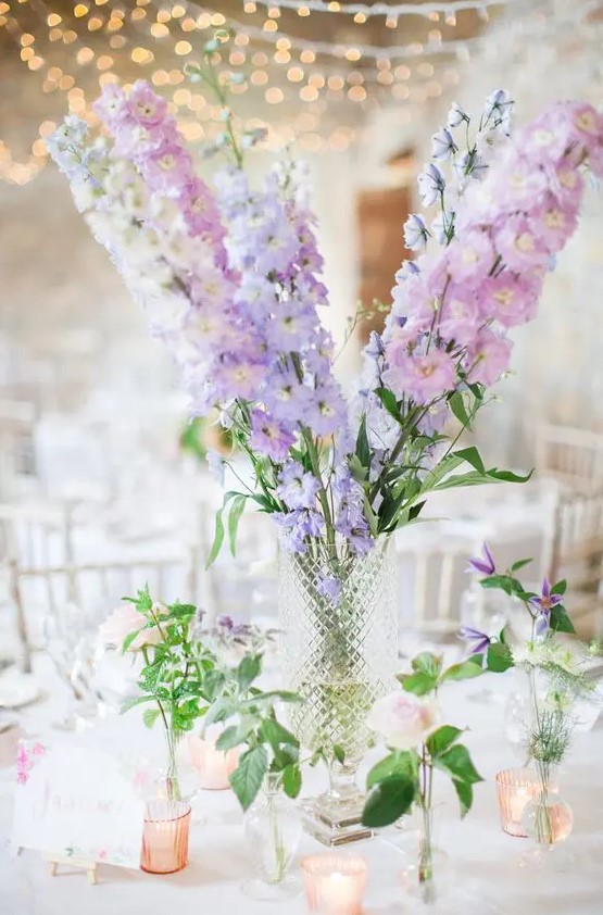 a lovely tall wedding centerpiece of white, lilac and blush delphinium and greenery in smaller vases around is cool