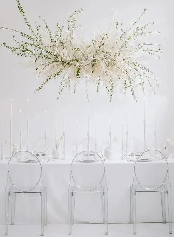 a minimalist ethereal wedding reception space with an oversized overhead floral decoration, sheer chairs and everything white