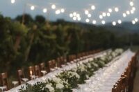 a neutral vineyard wedding reception with a greenery runner and white blooms, white linens plus string lights over the space
