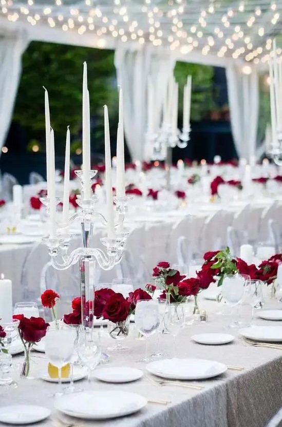 a neutral wedding tablescape with a shiny silver tablecloth, red roses, crystal candelabras and some greenery is a modern take on classics