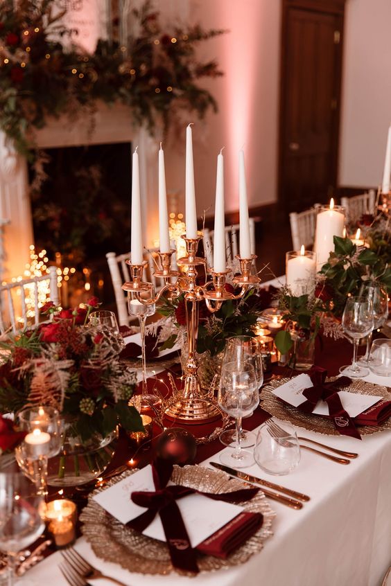 a refined NYE wedding tablescape with a burgundy table runner and napkins, ornaments, greenery, red roses and a candelabra with tall and thin candles