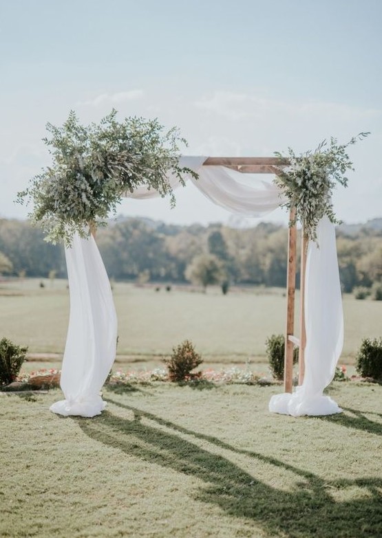 a simple rustic spring wedding arch with flowy white fabric and lots of greenery looks fresh and cool