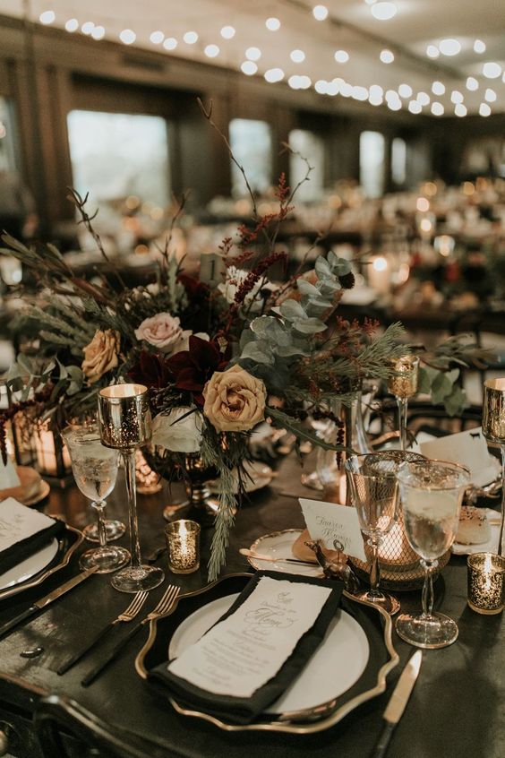 a sophisticated NYE wedding table setting with a black tablecloth, bold and neutral blooms and greenery, candles and gold rimmed glasses