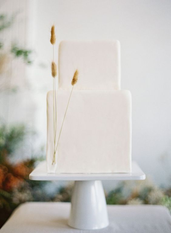 a white minimalist wedding cake of a square shape, decorated with just three twigs of dried grasses