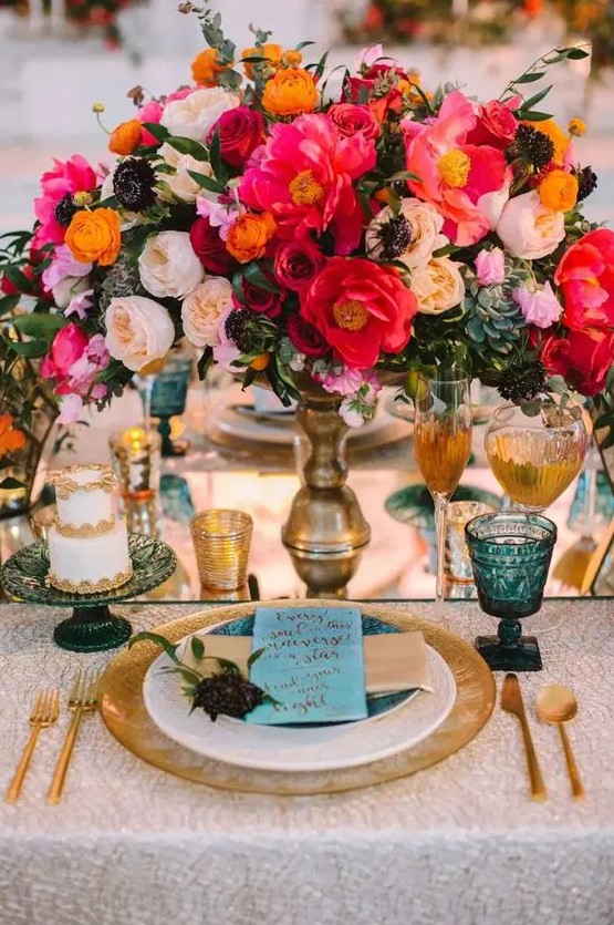 a bold jewel tone wedding table setting with pink, red, peachy and yellow blooms and succulents, blue glasses, gold touches
