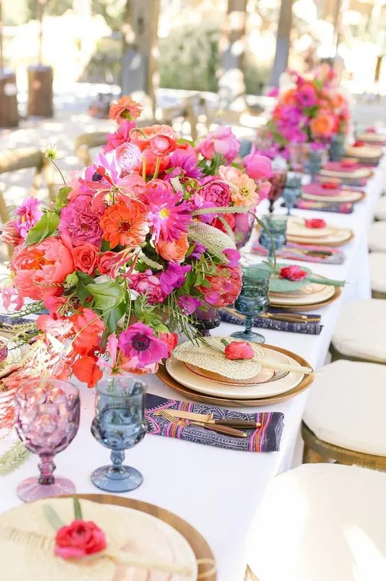a colorful wedding table setting with hot pink and red blooms, purple and blue glasses, navy and pink napkins and greenery