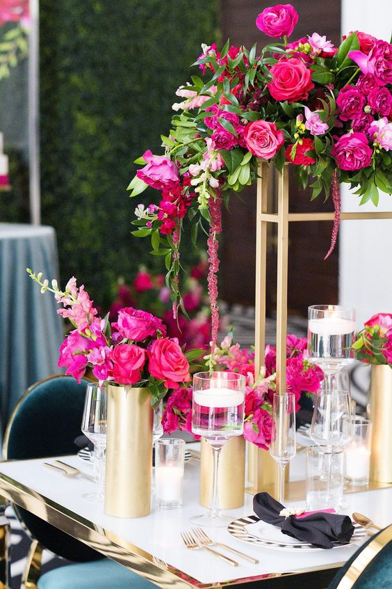 a bold wedding centerpiece on tall gold stands, with hot pink and fuchsia blooms and greenery plus matching arrangements on the table