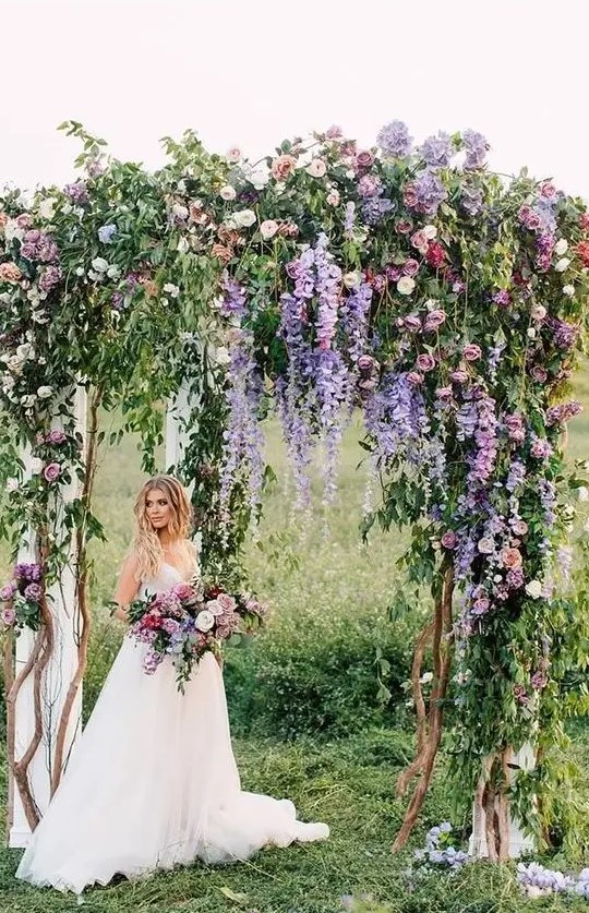 a very natural wedding arch with much greenery, purple and light pink blooms and some neutral fabric is wow