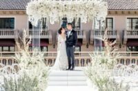 68 a gorgeous wedding ceremony space with a lucite wedding arch with lush white blooms, white blooms lining up the aisle and a glossy white floor