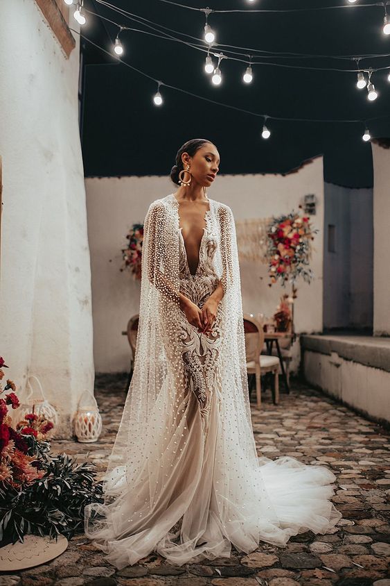 a jaw dropping lace mermaid wedding dress with a plunging neckline and a pearl and crystal long bridal capelet