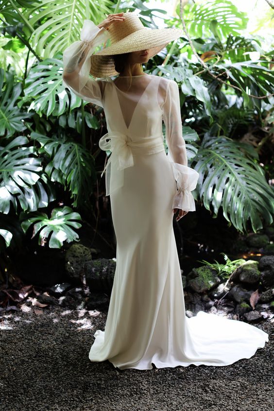 a minimalist plain wedding dress with a deep neckline, a train, with a sheer wrap dress topper with long sleeves and a hat