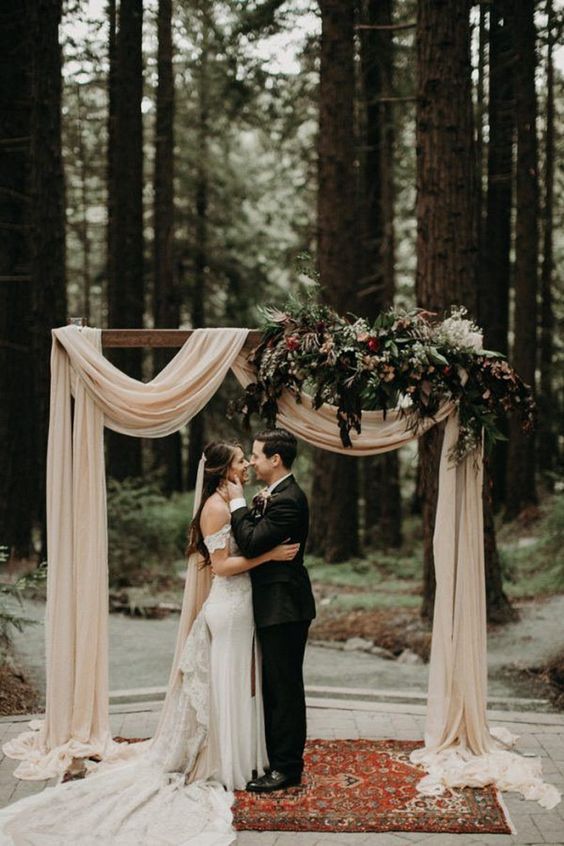 a beautiful and refined wedding arch with neutral drapes, greenery and dark blooms plus a boho rug is a stylish idea for a wedding