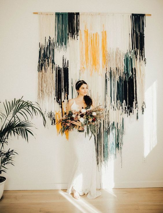 a boho wedding backdrop of white, yellow and dark green and black fringe plus greenery around is a lovely idea to try