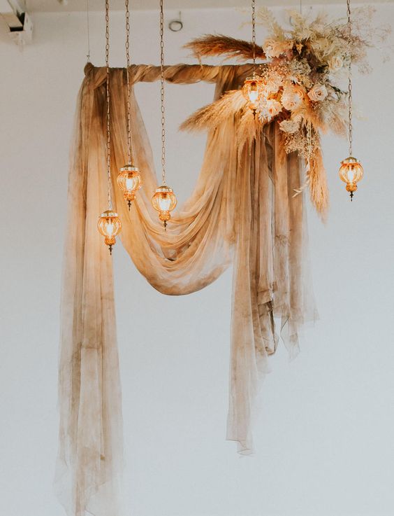 a beautiful drape wedding arch of watercolor fabric, some neutral blooms and pampas grass on the corner and hanging bulbs is wow
