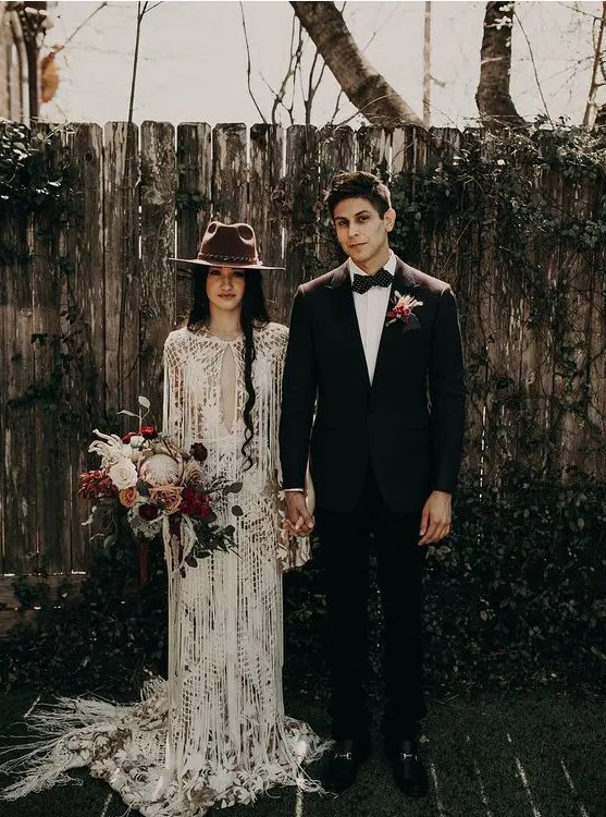 a boho lace sheath wedding gown with a plunging neckline, a train and long fringe plus a brown hat