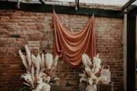 08 a boho wedding backdrop with a rust-colored draped, pampas grass and woven lanterns plus a bold boho rug