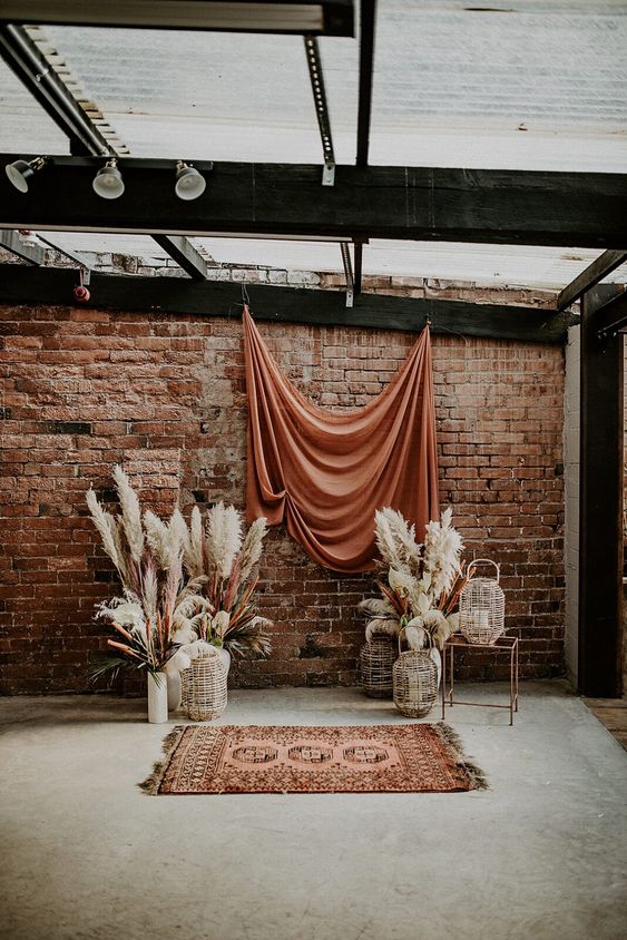 a boho wedding backdrop with a rust colored draped, pampas grass and woven lanterns plus a bold boho rug