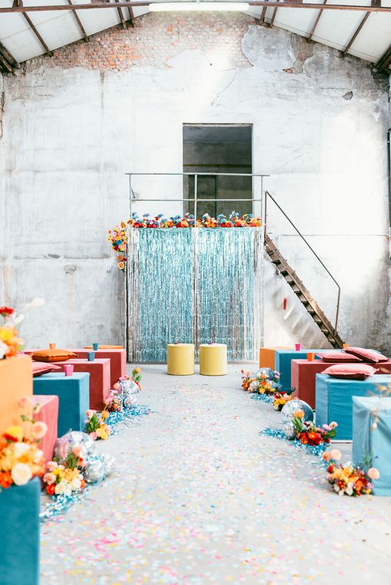 a colorful wedding ceremony space with a turquoise tinsel wedding backdrop, colorful poufs and stools and bright blooms