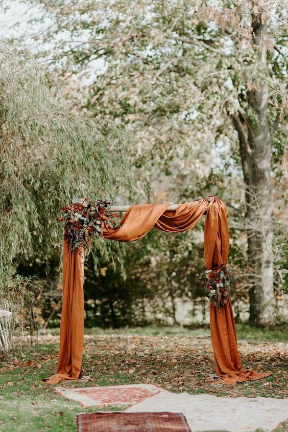 a bold fall wedding arch with rust colored drapes, greenery, white and burgundy blooms and boho rugs on the ground