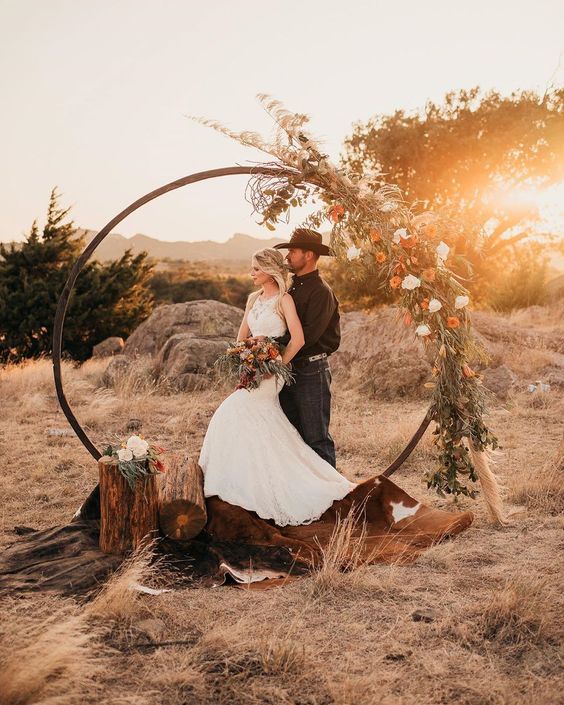 a western wedding arch with greenery, white and orange blooms, a cowhide rug, tree stumps is a perfect idea for a boho ceremony