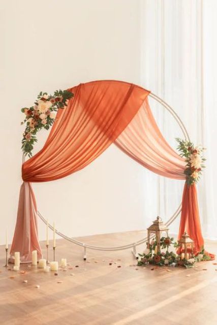 a bright wedding backdrop of a large hoop, orange drapes, neutral and orange blooms, candles and candle lanterns around