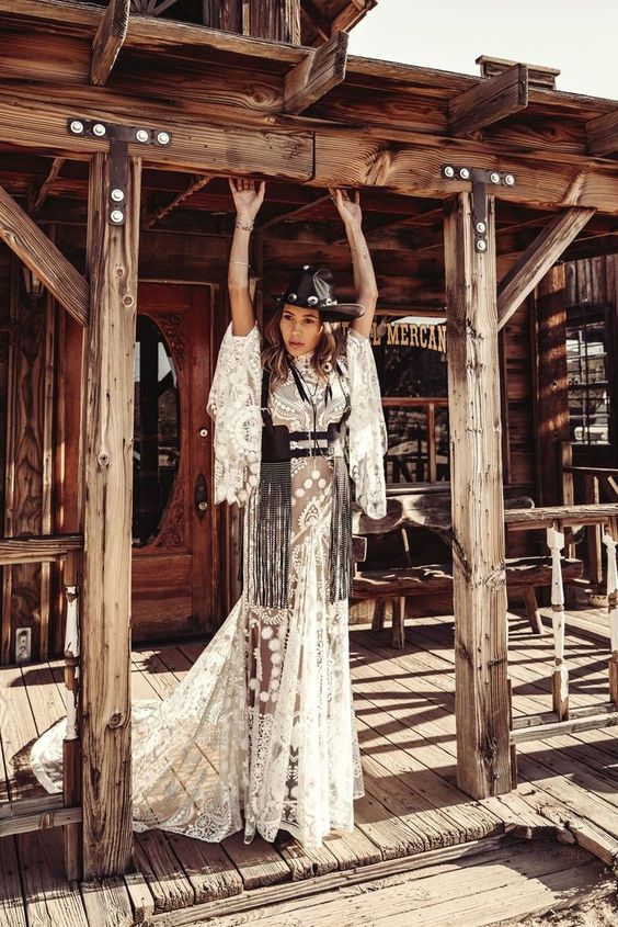 a catchy western bridal look with a lace applique wedding dress with bell sleeves and a train, a black leather corset with long fringe and a black embellished hat