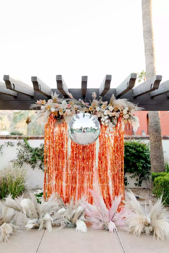 a fun wedding backdrop of orange tinsel fringe, a disco ball, pampas grass and neutral blooms is a fantastic solution