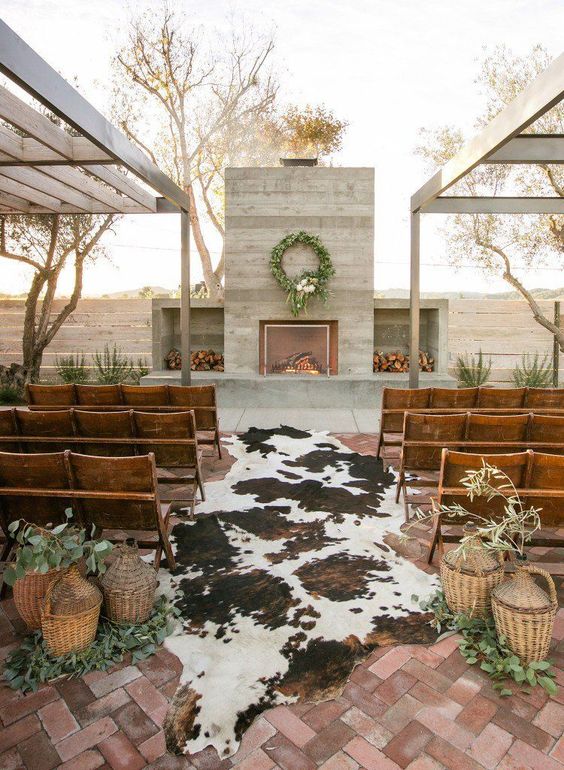 a western wedding ceremony space with a fireplace, firewood storage, cowhides, benches, baskets and greenery is welcoming
