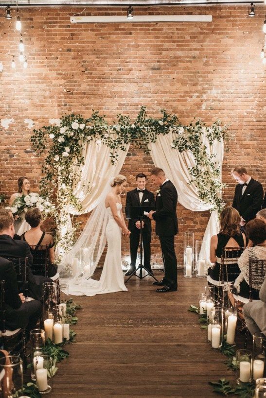 a chic wedding backdrop of white drapes, greenery and white blooms and pillar candles in tall glasses is a lovely idea for a wedding