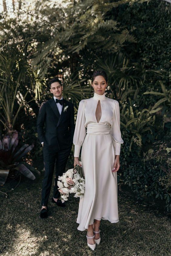 a modern A-line midi wedding dress with a turtleneck, draped sleeves and a waistline, white shoes are a lovely modern bridal look
